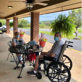 Two individuals enjoying a sunny day outside, one seated in a wheelchair, both wearing masks.