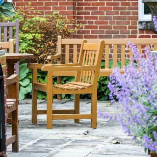 A tranquil garden patio with wooden chairs and benches surrounded by lush plants and flowers.
