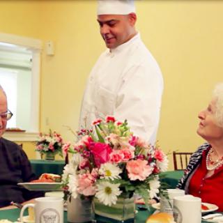 A waiter serves a meal to an elderly couple at a beautifully arranged dining table adorned with flowers.