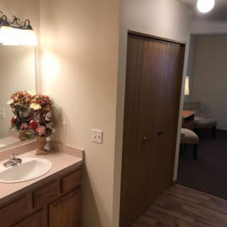 A small bathroom with a sink and mirror, leading to a hallway with visible furniture.
