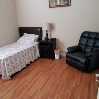 A simple bedroom with a single bed, nightstand, and recliner chair on hardwood flooring.