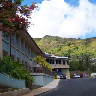 A scenic view of a building nestled against a mountainous backdrop with a parking lot in the foreground.