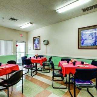 A small dining area with tables covered in red tablecloths and chairs arranged around them. Paintings adorn the walls, and fluorescent lights illuminate the room.