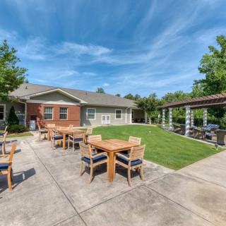 A sunny patio area with tables and chairs, adjacent to a house and lawn.