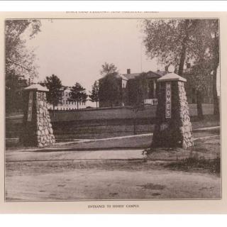 A historical photograph of a stone gate entrance, featuring two large pillars and a dirt road leading to a distant building surrounded by trees.