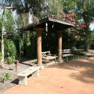 A tranquil garden scene featuring a shaded pergola with benches and surrounding greenery.
