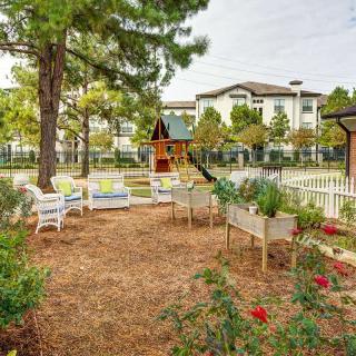 A serene garden area with chairs and a playground, surrounded by trees and overlooking a building.