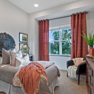 A cozy bedroom with warm-toned decor, featuring a bed with pillows and blanket, and a window with red curtains.