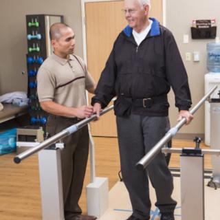 An elderly man receiving assistance from a physical therapist while walking with parallel bars in a rehabilitation center.