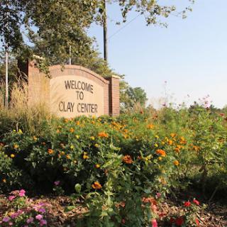 A welcoming sign to Clay Center, surrounded by vibrant flowers and lush greenery.