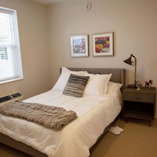A modern bedroom with a neatly made bed, gray headboard, and decorative pillows, complemented by wall art and a nightstand with a lamp.