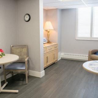 A cozy living room with gray walls, wooden flooring, and minimalistic furniture including a table, chairs, and a lamp.
