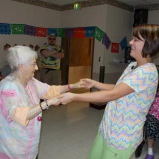 An elderly woman and her caregiver share a joyful dance at an indoor celebration.