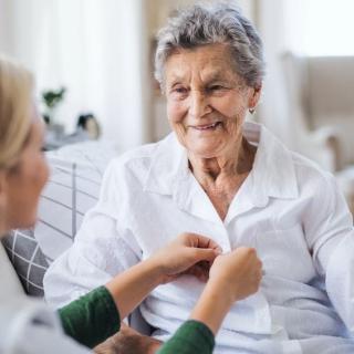 A caregiver helping an elderly woman button her shirt while they both smile.