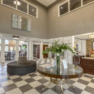 A spacious lobby area with a checkered floor, featuring a central round table adorned with flowers and surrounded by various pieces of furniture.