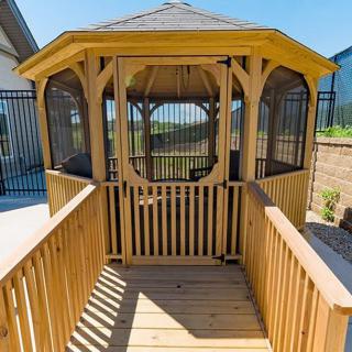 A wooden gazebo with a walkway leading to the entrance, surrounded by fencing on a sunny day.