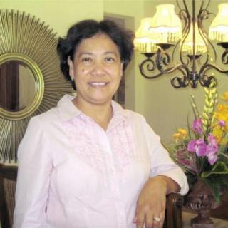 A woman standing indoors, smiling beside a decorative table with flowers and a mirror in the background.
