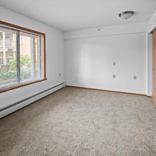 A spacious empty room with natural light streaming in through the window, featuring neutral-colored walls and a carpeted floor.