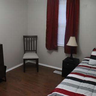 A simple bedroom with a bed, chair, and lamp next to a window with red curtains.