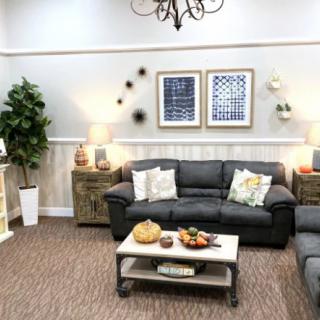 A cozy living room featuring a black leather sofa, decorative pillows, a wooden coffee table with fruit and decor items, framed artwork on the walls, ambient lighting, and a potted plant.