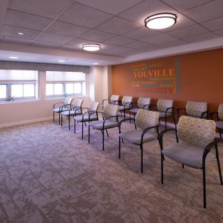 An indoor waiting room with several chairs arranged in rows, featuring windows and carpeted flooring.