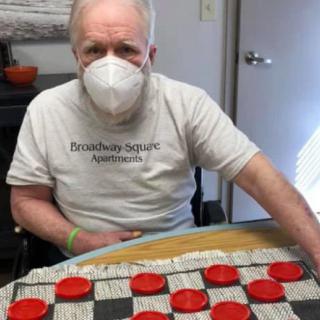 An elderly man wearing a mask is sitting at a table playing checkers indoors.