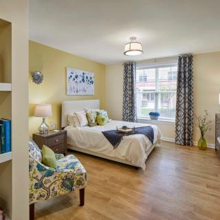A modern and well-decorated bedroom with wood flooring, a comfortable bed, stylish chairs, and ample natural lighting.