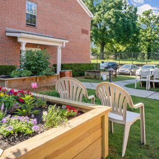 A cozy outdoor garden area with chairs and a flower bed next to a brick building on a sunny day.