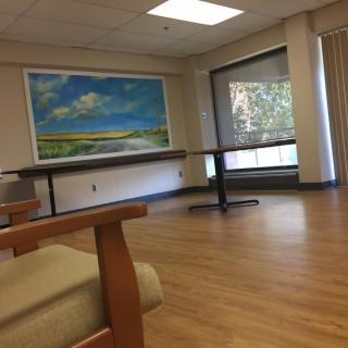 A spacious room with wooden flooring, decorated with a landscape painting and large windows.