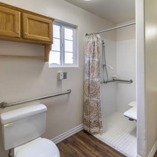 A bathroom featuring a shower with grab bars, a toilet, and a wooden cabinet with a window above.
