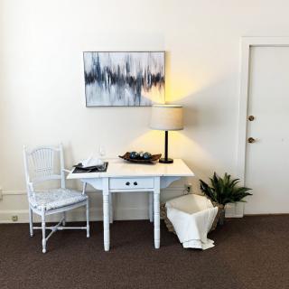 A minimalist room setup with a white table and chair, accompanied by a lamp, artwork, and plant.