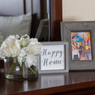 A cozy corner featuring a framed 'Happy Home' sign, white flowers in a vase, and a colorful photo on a wooden surface.
