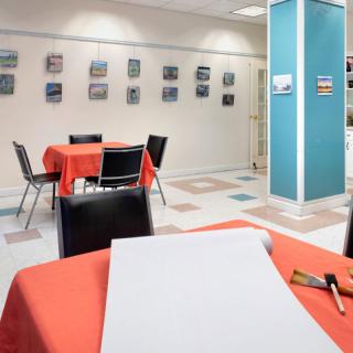 An interior view of an art gallery with tables and chairs, displaying various artworks on the wall.