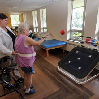 An elderly person engages in a physical therapy session, assisted by a caregiver, practicing throwing skills indoors.