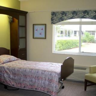 A simple room featuring a hospital bed near a window with an armchair and floral curtains.