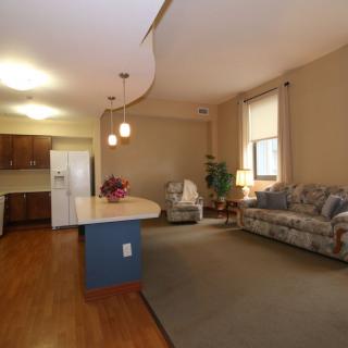 An open floor plan living area with a kitchen in the background, featuring hardwood floors, a cozy sofa, and warm lighting.
