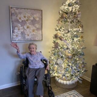 An elderly person in a wheelchair waves beside a beautifully decorated Christmas tree in a cozy living room setting.