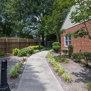 A serene garden pathway alongside a brick building with lush greenery and trees.