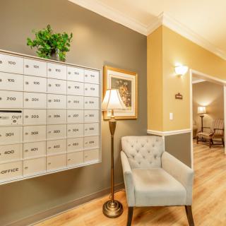 A cozy hallway featuring a set of mailboxes, a comfortable chair, a standing lamp, and decorative elements such as a painting and a plant.