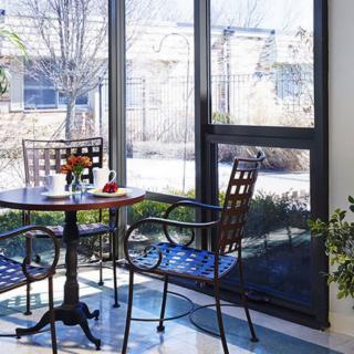A sunlit room with a table and chairs by large windows, surrounded by plants.