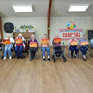 A group of seniors holding paintings seated indoors at a carnival-themed event.