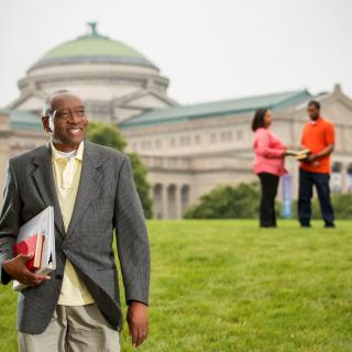 A man holding books smiles while standing on a grassy area with a large building in the background.