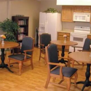 A cozy kitchen area with wooden tables and chairs, featuring a bookshelf, indoor plant, and standard kitchen appliances.