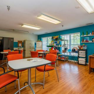 A well-lit classroom with tables and orange chairs, featuring bookshelves and educational materials.