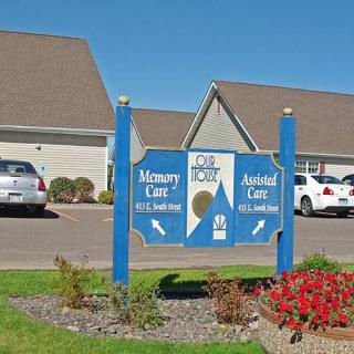 A sign for a memory care and assisted living facility in front of buildings and parked cars.