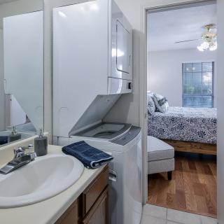 A compact bathroom features a laundry area with a washer and dryer, next to a sink and mirror, leading into a cozy bedroom.