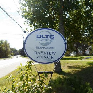 A sign for DLTC Healthcare's Bayview Manor is displayed by the roadside, surrounded by trees.