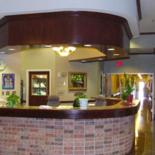 A reception desk in a well-lit office lobby with plants and a corridor leading further inside.