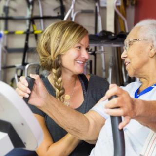 A caregiver assisting an elderly person on exercise equipment.