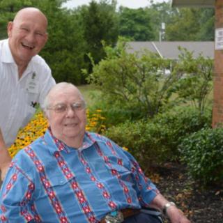 Two people enjoying time outside in a garden, one sitting in a wheelchair while the other stands nearby.